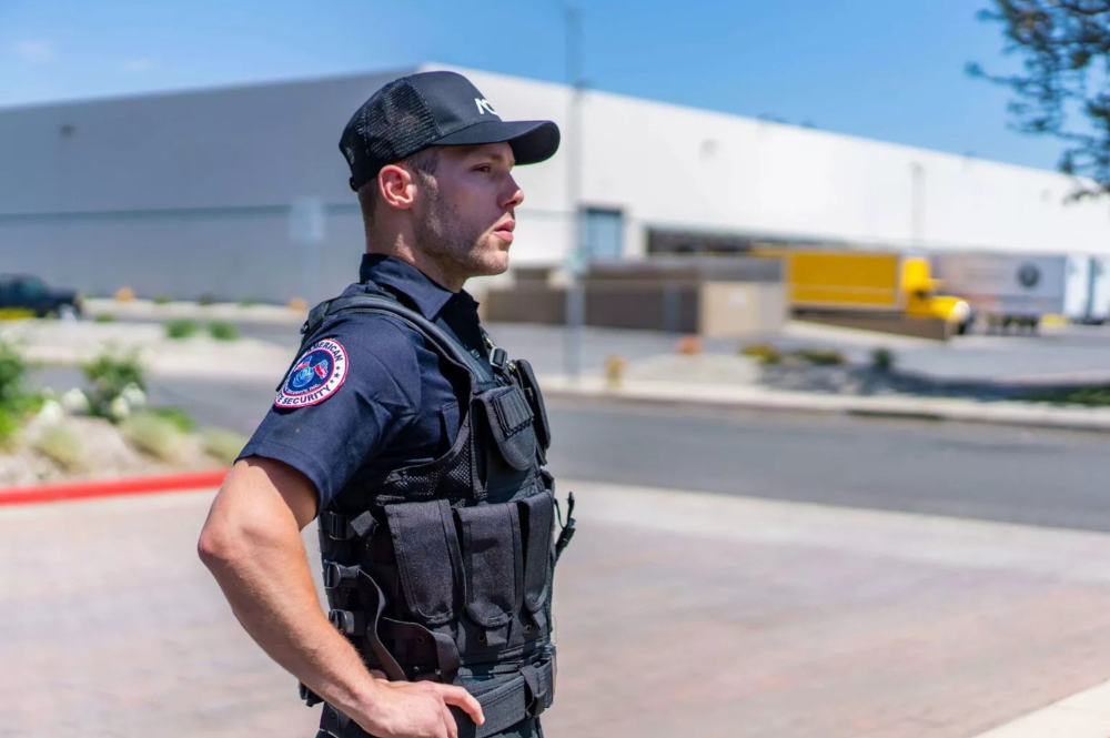 Uniformed security guard in a bullet-proof vest and black cap standing outside a large industrial building, looking attentively to the side.