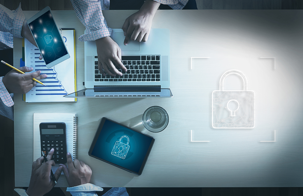Overhead view of hands typing on a laptop and holding a tablet on a tabletop, overlaid with a semi-transparent padlock icon representing cybersecurity.